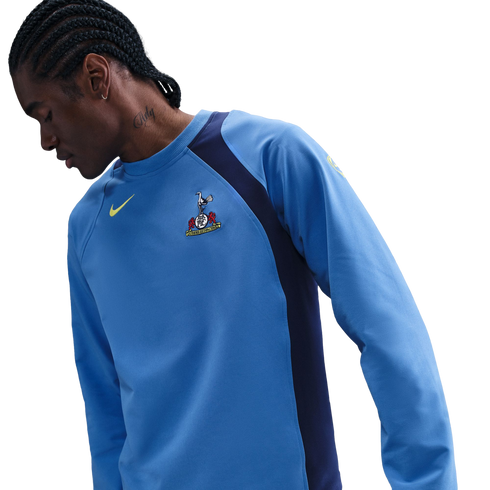 A man with braided hair and a neck tattoo wears a light blue Nike Tottenham Hotspur training shirt with navy blue side panels, looking down against a black background.