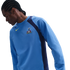 A man with braided hair and a neck tattoo wears a light blue Nike Tottenham Hotspur training shirt with navy blue side panels, looking down against a black background.