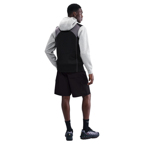 Rear view of a man in a grey hoodie, black vest, black cargo shorts, and sneakers against a black background.