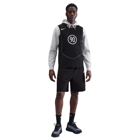 A Black man in a gray hoodie, black Nike jersey with "90" logo, black cargo shorts, and sneakers, standing against a solid black background.