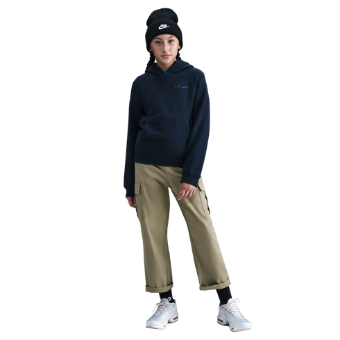 A person wearing a black Nike beanie, navy Nike "CFC" hoodie, tan cargo pants, and light blue sneakers, standing against a black background.