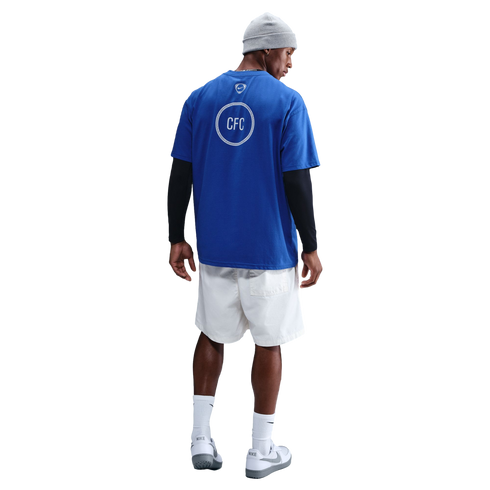 Rear view of a man wearing a blue CFC t-shirt over black long sleeves, white shorts, a grey beanie, and Nike sneakers against a black background.