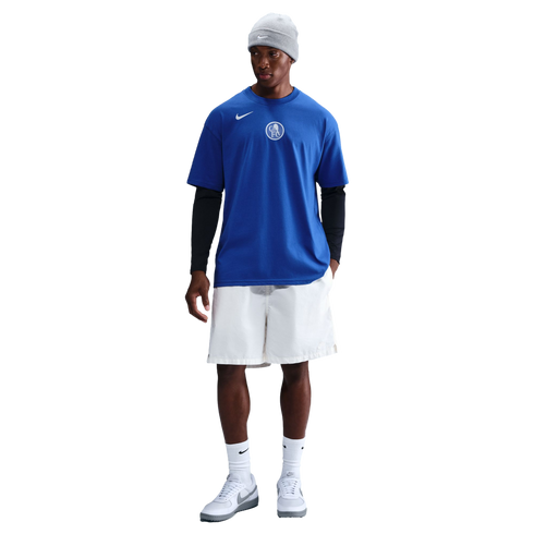 A person in a blue Nike Chelsea FC t-shirt over black long sleeves, white shorts, and a grey beanie stands against a black background.
