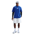 A person in a blue Nike Chelsea FC t-shirt over black long sleeves, white shorts, and a grey beanie stands against a black background.