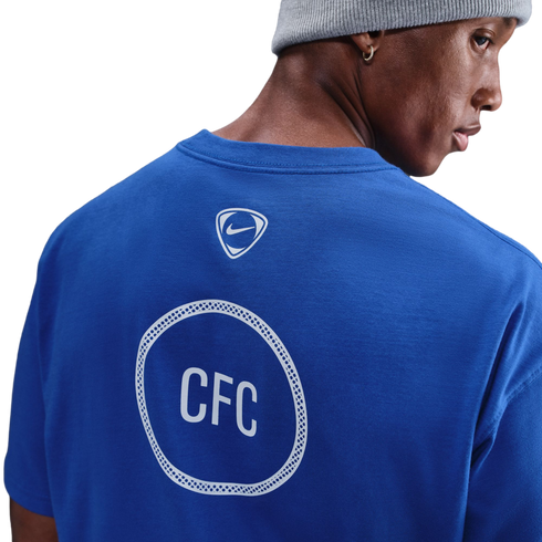 Back view of a man in a blue Chelsea FC t-shirt and grey beanie, showing a Nike logo and "CFC" inside a white patterned circle on the shirt.