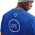 Back view of a man in a blue Chelsea FC t-shirt and grey beanie, showing a Nike logo and "CFC" inside a white patterned circle on the shirt.