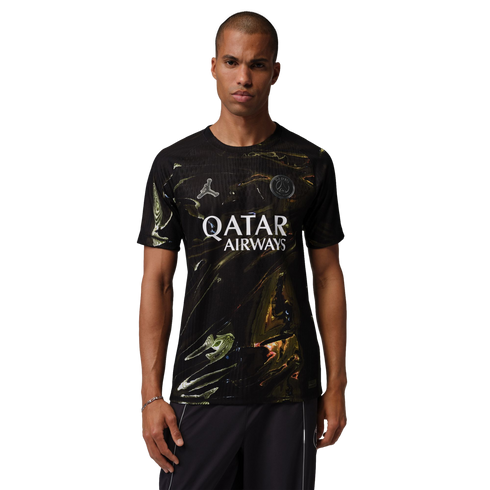 A man models a black Paris Saint-Germain soccer jersey featuring a gold and green marbled pattern. The jersey displays a silver Jordan Jumpman logo, the club crest, and "QATAR AIRWAYS" in white across the chest. He is standing against a solid black backgr