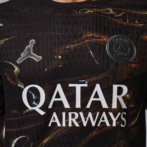 A close-up of a black Paris Saint-Germain soccer jersey with a brown and gold marble-like pattern. It features the white Jordan Brand "Jumpman" logo on the right chest, a black and silver PSG crest on the left chest, and the "QATAR AIRWAYS" sponsor logo i