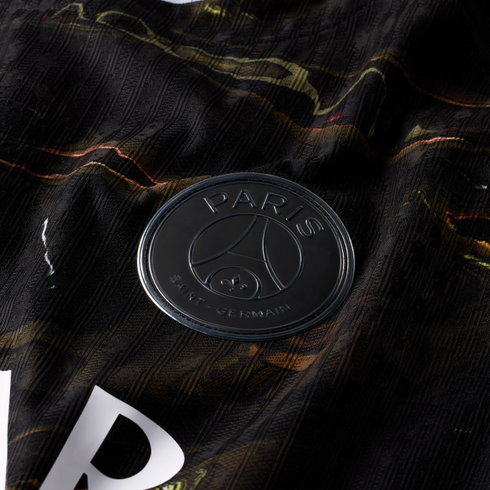 A close-up photograph of a matte black Paris Saint-Germain (PSG) football club badge. The circular badge, featuring raised lettering and the team's iconic Eiffel Tower and fleur-de-lis logo, is set against a dark, ribbed fabric jersey with marbled gold an
