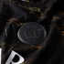 A close-up photograph of a matte black Paris Saint-Germain (PSG) football club badge. The circular badge, featuring raised lettering and the team's iconic Eiffel Tower and fleur-de-lis logo, is set against a dark, ribbed fabric jersey with marbled gold an