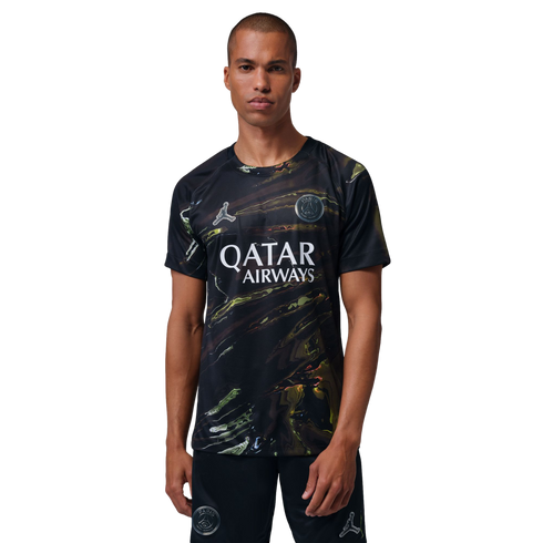 A man with a buzz cut models a black Paris Saint-Germain jersey and shorts featuring an iridescent marbled pattern, the Qatar Airways sponsor logo, and Jordan Brand's Jumpman logo, set against a solid black background.