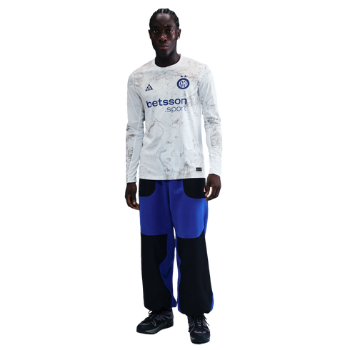 A young Black man with braids wears a long-sleeve white jersey with gray patterns and a "betsson .sport" logo, blue and black pants, and dark sneakers, standing against a black background.