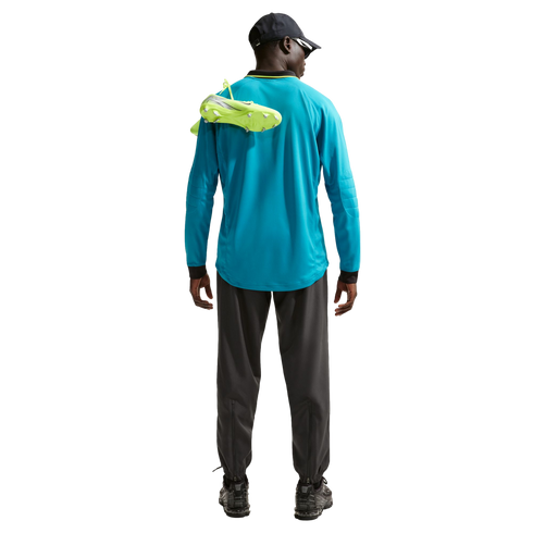 Rear view of a person in a blue long-sleeved shirt and grey trousers, carrying neon yellow soccer cleats over their shoulder against a black background.