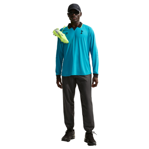A person in a blue Tottenham long-sleeve polo, grey joggers, black cap, and sunglasses with neon green soccer cleats draped over their shoulder.