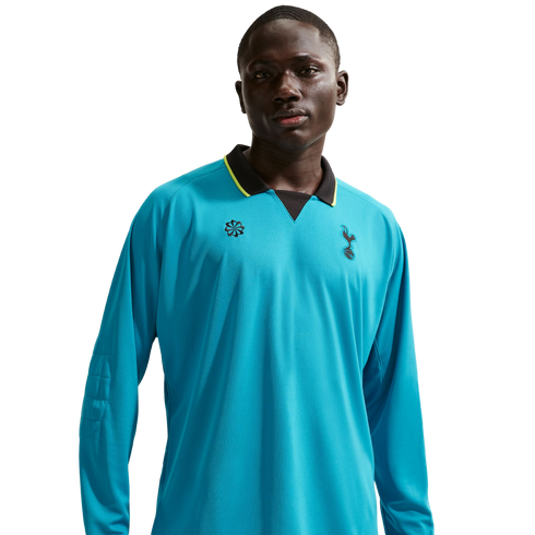 A man wearing a long-sleeved, bright blue Tottenham Hotspur soccer jersey with a black and yellow collar, set against a black background.