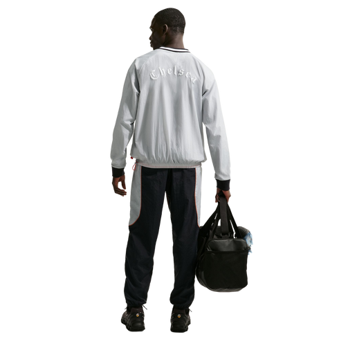 Rear view of a man in a grey 'Chelsea' jacket and navy track pants, holding a black duffel bag against a black background.