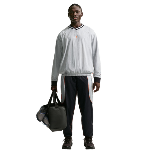 Full-length shot of a man in a grey pullover and navy track pants, holding a black duffel bag with blue cleats against a black background.