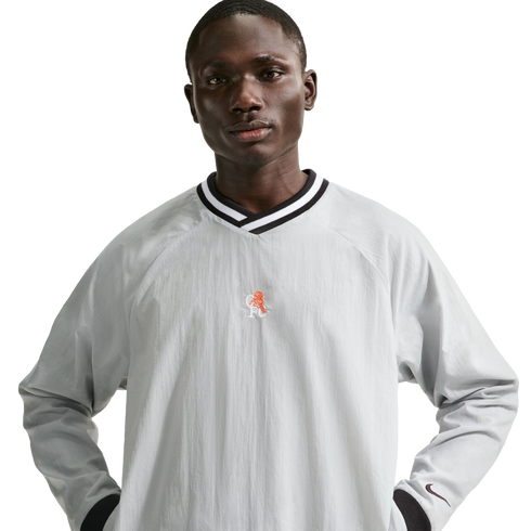 A man in a light grey Nike windbreaker shirt with a striped v-neck and orange lion crest, posing against a black background.