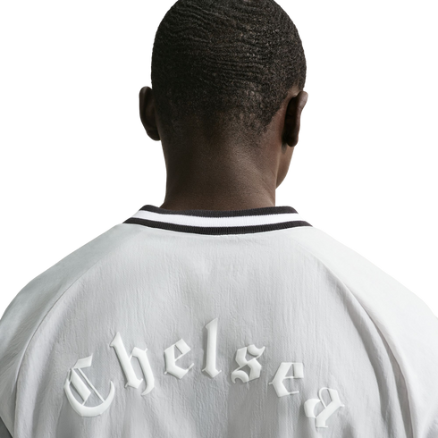 View from behind of a person wearing a white jacket. Across the upper back, the word "Chelsea" is embossed in a white, gothic-style font. The jacket has a black and white striped collar.