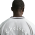 View from behind of a person wearing a white jacket. Across the upper back, the word "Chelsea" is embossed in a white, gothic-style font. The jacket has a black and white striped collar.