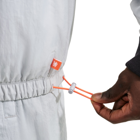 Close-up of a hand pulling an orange drawstring toggle on a light gray elastic waist garment with a small orange logo tag.