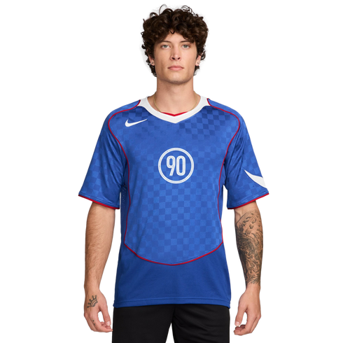 A man with curly hair wearing a blue Nike Total 90 soccer jersey with a white "90" logo and checkered pattern.