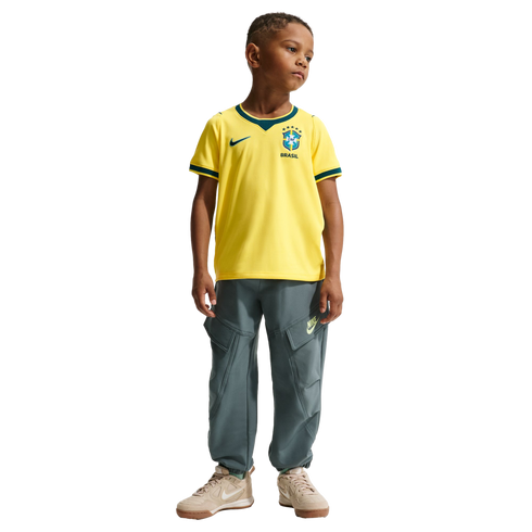 A full-body studio portrait of a young boy standing against a black background. He is wearing a yellow Nike Brazil soccer jersey with green trim, grey cargo-style jogger pants, and tan sneakers.