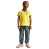 A full-body studio portrait of a young boy standing against a black background. He is wearing a yellow Nike Brazil soccer jersey with green trim, grey cargo-style jogger pants, and tan sneakers.
