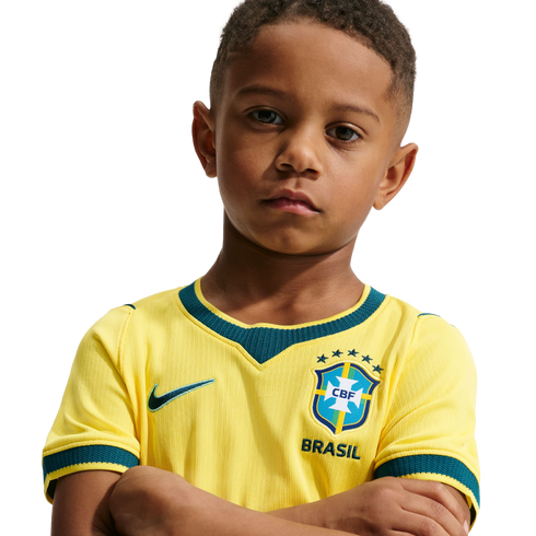 A young boy with a serious expression and arms crossed, wearing a yellow Brazil national soccer team jersey against a solid black background.