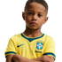 A young boy with a serious expression and arms crossed, wearing a yellow Brazil national soccer team jersey against a solid black background.
