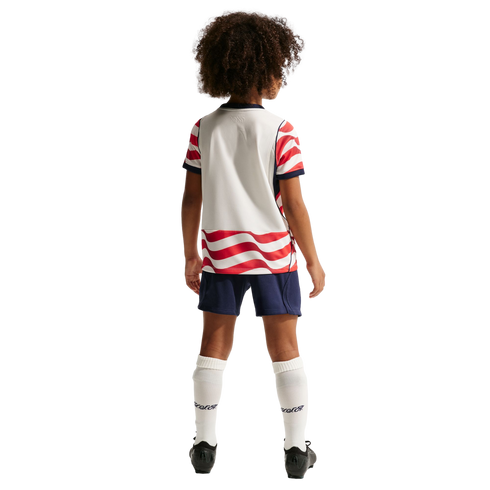 Back view of a child with curly hair wearing a white and red wavy-striped soccer uniform, blue shorts, and white socks against a black background.