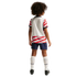 Back view of a child with curly hair wearing a white and red wavy-striped soccer uniform, blue shorts, and white socks against a black background.