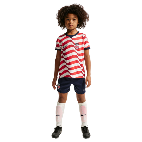 Young child with curly hair wearing a red and white wavy-striped US soccer jersey, navy shorts, and white socks against a solid black background.