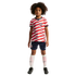 Young child with curly hair wearing a red and white wavy-striped US soccer jersey, navy shorts, and white socks against a solid black background.