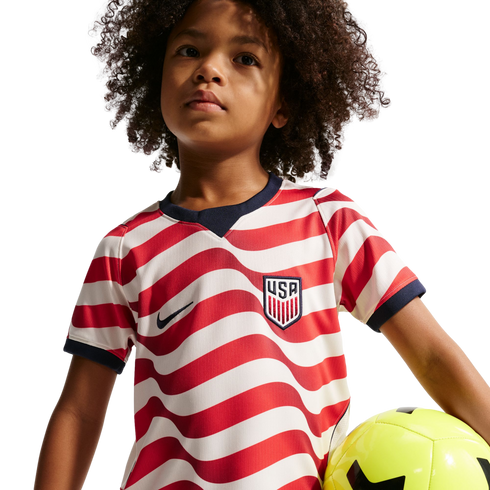 A young child with curly hair wearing a red and white striped USA soccer jersey holding a yellow soccer ball against a black background.