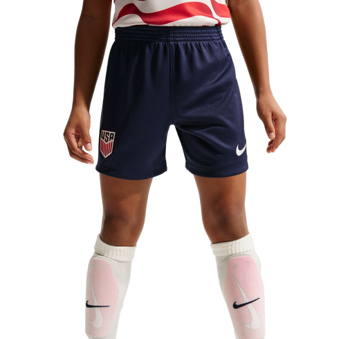 A child wears navy blue USA soccer shorts with a Nike logo, a red and white jersey, and white Nike socks with pink shin guards against a black background.