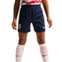 A child wears navy blue USA soccer shorts with a Nike logo, a red and white jersey, and white Nike socks with pink shin guards against a black background.