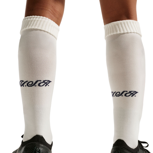 A close-up of a person's legs wearing white knee-high socks with a navy blue "USA" script logo on the calves and black athletic shoes, set against a solid black background.