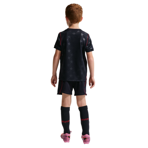 Rear view of a young boy with red hair wearing a black soccer kit with a star pattern and red trim. He is standing against a solid black background, wearing tall black socks with "U.S.A." in red and pink soccer cleats.
