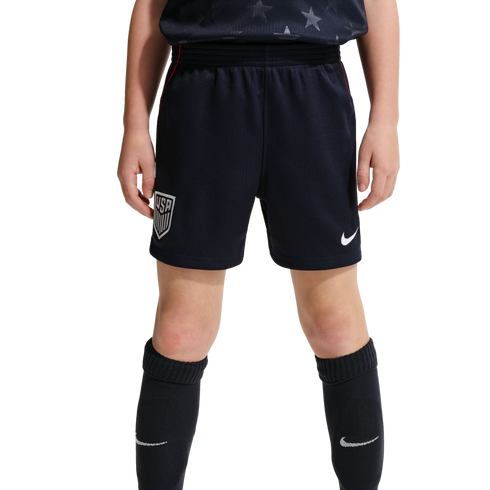 A front-facing view of a youth athlete from the waist down wearing navy blue USA national team soccer shorts and matching knee-high Nike socks against a black background.