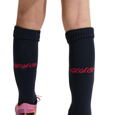A close-up of a person's lower legs against a black background, wearing navy blue knee-high socks with a red script logo and a glimpse of a pink and orange athletic shoe.