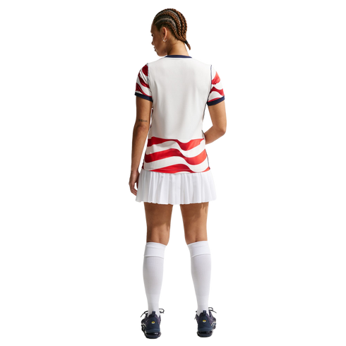 Rear view of a person in a white sports jersey with red wavy stripes, a white pleated skirt, and knee-high socks against a black background.