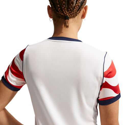 Back view of a person with braided hair wearing a white sports jersey with red and white patterned sleeves and navy blue trim.
