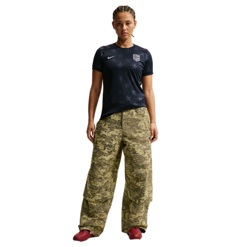 A woman stands against a black background wearing a navy USA soccer jersey, oversized camouflage cargo pants, and red sneakers.