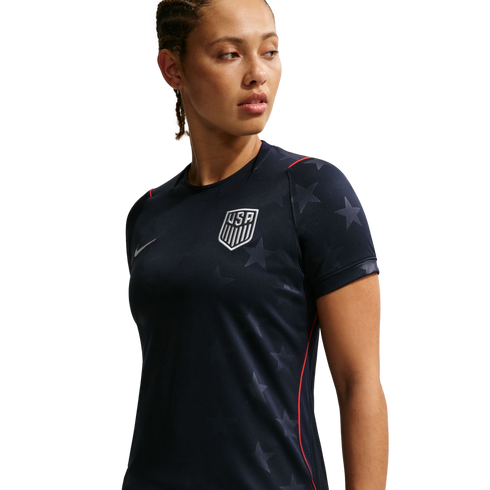 A woman in a navy blue USA soccer jersey with subtle star patterns and a white team crest, looking to her right against a black background.