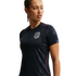 A woman in a navy blue USA soccer jersey with subtle star patterns and a white team crest, looking to her right against a black background.
