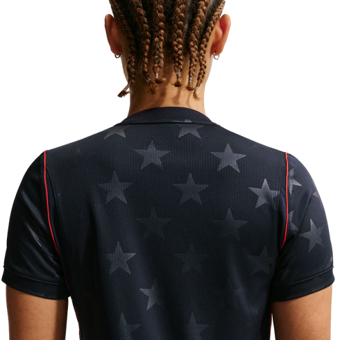Back view of a person with brown braided hair wearing a navy blue shirt with a grey star pattern and red trim on the sleeves, against a black background.