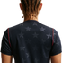 Back view of a person with brown braided hair wearing a navy blue shirt with a grey star pattern and red trim on the sleeves, against a black background.