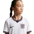 A young girl in a white England national football team jersey looking to the side against a black background.
