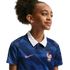 A young girl in a blue patterned France national team soccer jersey with a white collar, looking off-camera against a black background.